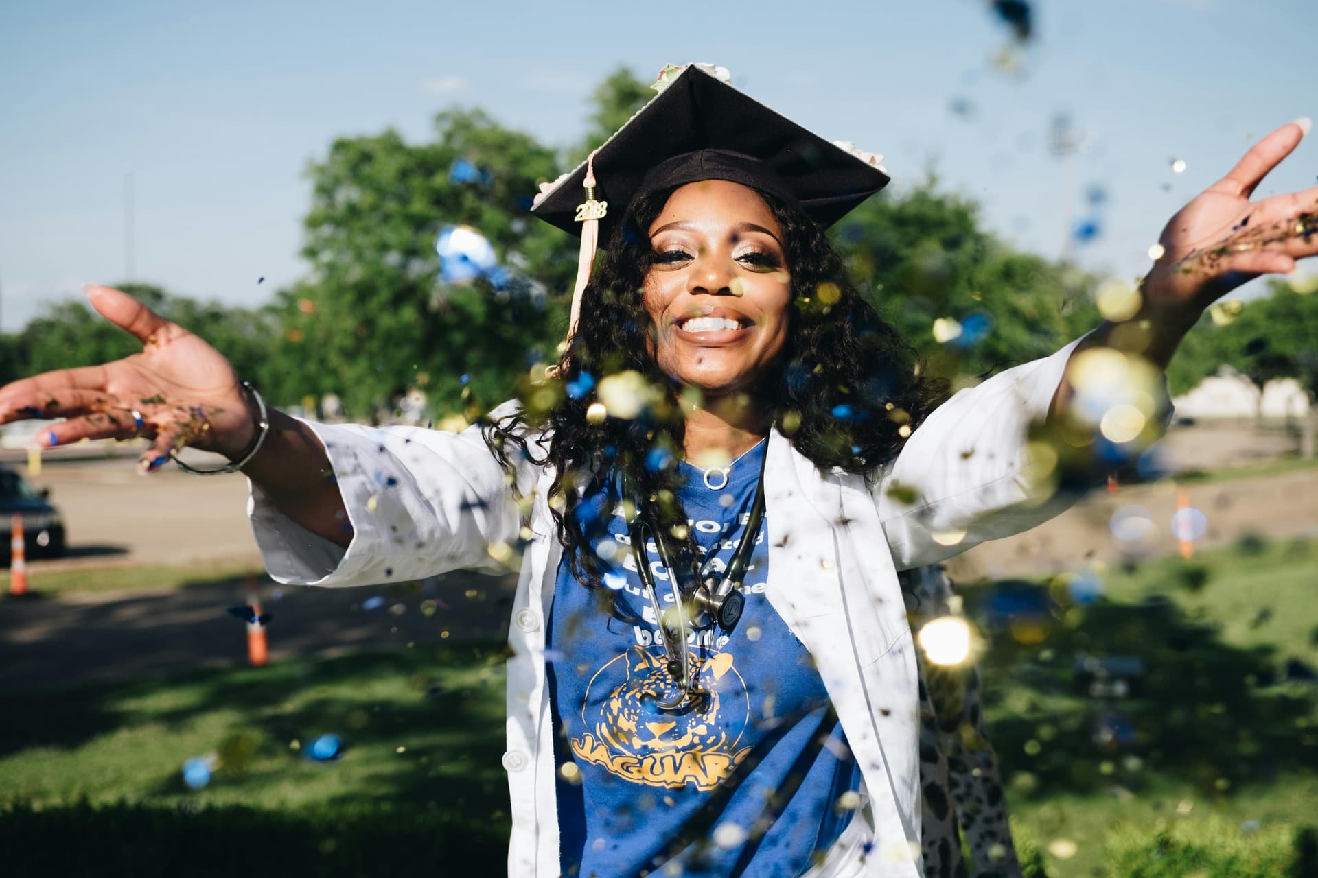 A person wearing a graduation cap and a white coat joyfully throws confetti outdoors, celebrating their achievement.