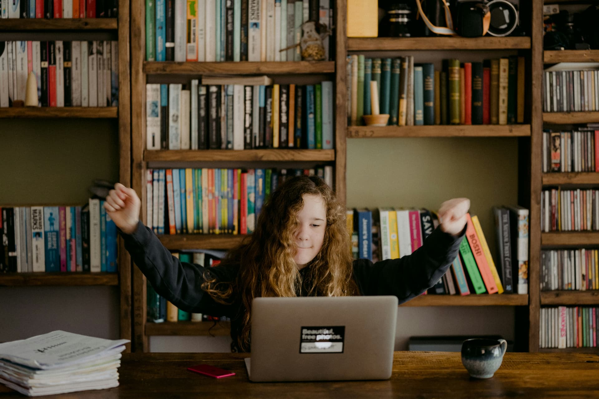 Girl with long hair raising arms in front of laptop, bookshelves behind.